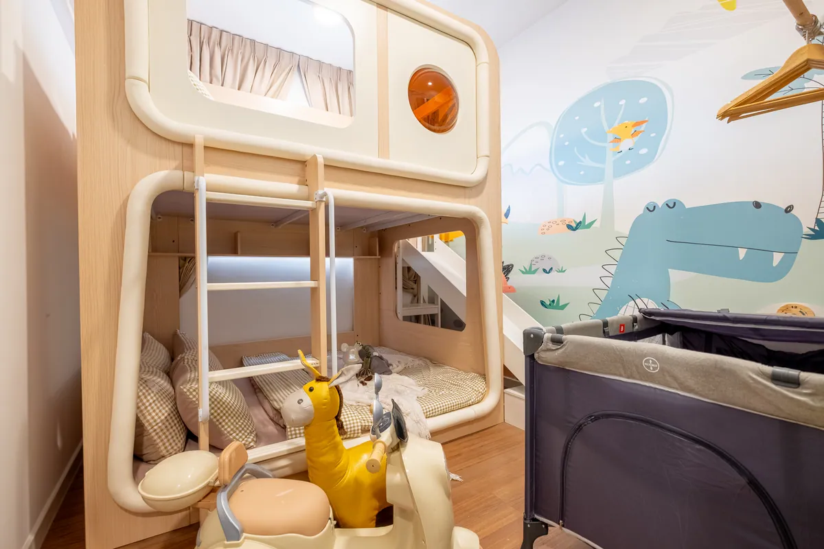 The kids' room at Kidtastic Stay KL: a bunk house with a round window, a dinosaur mural, ride-on toys and a baby cot.
